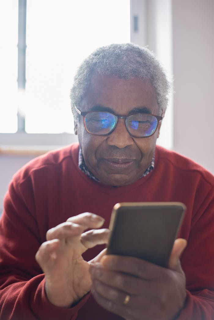 Elderly man with gray hair using a smartphone, sitting indoors with eyeglasses.