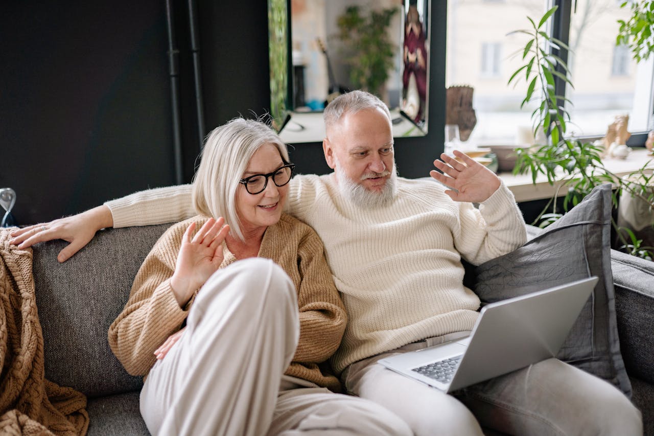 Happy senior couple in cozy sweaters video chatting on the sofa using a laptop. Indoor setting with natural light.
