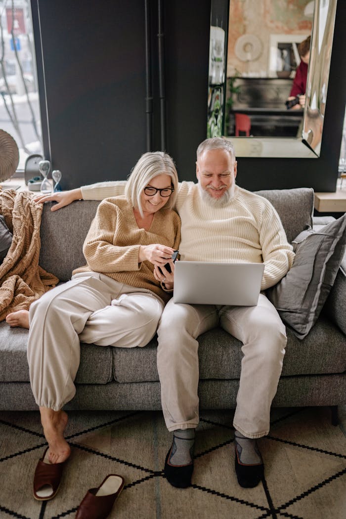 An elderly couple sits together on a couch sharing a laptop in a cozy indoor setting.