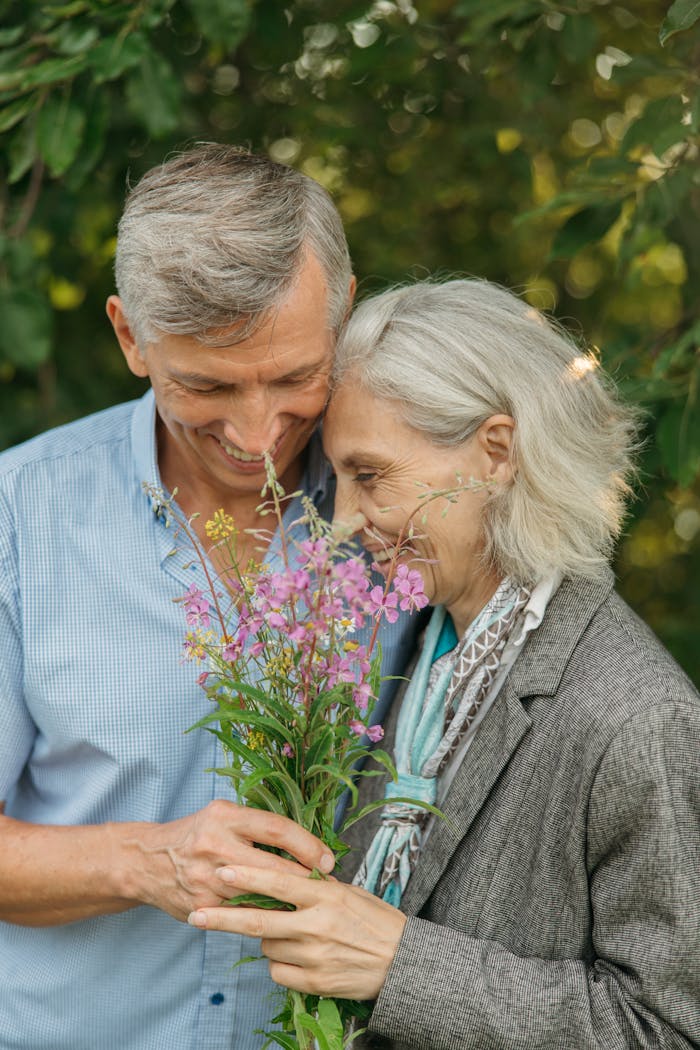 An elderly couple shares a joyful moment embracing flowers in an outdoor setting.
